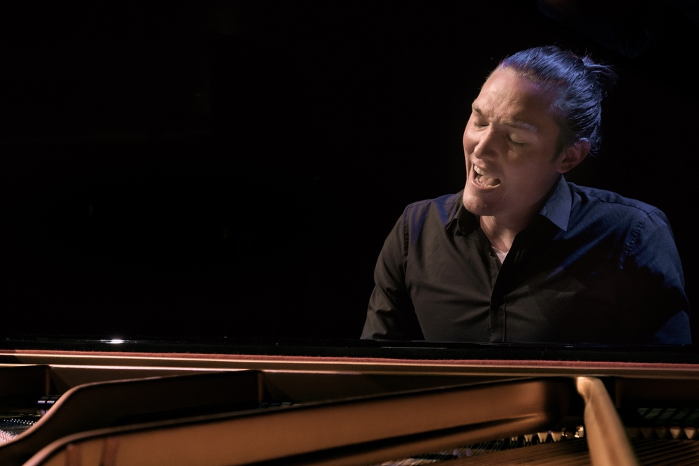 Andreas Langsch, Klavierkabarettist. Sitzt innig singend am Flügel vor schwarzem Hintergrund,