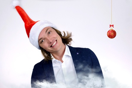 Andreas Langsch, Klavierkabarettists. Trägt einen Anzug, hat eine rote Nikolausmütze auf, neigt den Kopf lachend nach rechts. Links von ihm baumelt eine Weihnachtskugel.