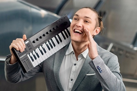 Andreas Langsch, Klavierkabarettist. Der Künstler steht auf einer Rolltreppe, hat ein kleines Keyboard rechts geschultert und lacht fröhlich.